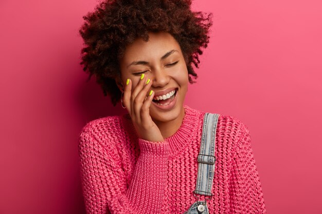 Concept d'émotions et de sentiments heureux. Joyeuse femme afro-américaine frisée rit d'entendre une blague hilarante, sourit largement, se divertir par un ami drôle, habillé avec désinvolture, pose à l'intérieur