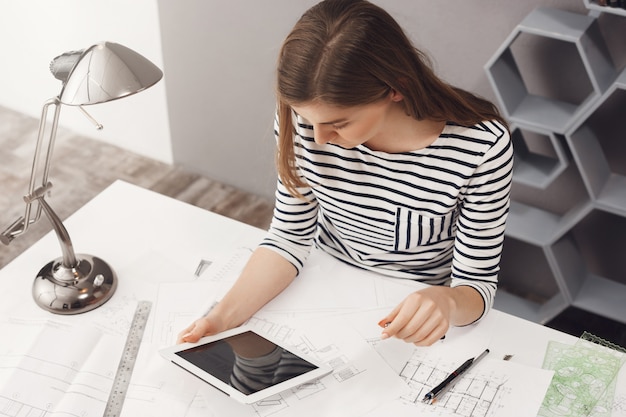 Concept d'emploi, de carrière et d'entreprise. Portrait de jeune designer féminin à la mode assis à table, à la recherche dans le moniteur de la tablette numérique, discutant avec le client pour décider de certains détails.