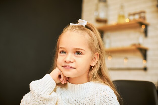 Concept de l'enfance. Image intérieure de la belle petite fille aux cheveux longs assis dans la cuisine avec guirlande portant un ruban blanc et pull tricoté, tenant la main sous son menton, souriant