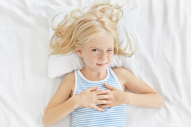 Concept d'enfants, de repos et de personnes. Jolie petite fille aux longs cheveux blonds, fermant un œil tout en voulant dormir, allongée dans un lit blanc va dormir. Fille de taches de rousseur au repos à la maison dans la chambre