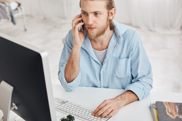 Concept d'entreprise, de bureau et de technologie. Vue de dessus d'un employé barbu portant une chemise bleue, parler au téléphone avec des compagnons, taper sur le clavier, regarder sur l'écran de l'ordinateur, à l'aide d'appareils modernes