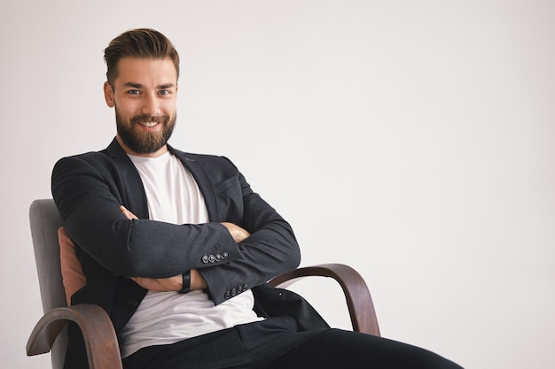 Concept d'entreprise, de carrière et de réussite. Enthousiaste élégant jeune homme d'affaires mal rasé avec un sourire heureux, assis dans un fauteuil contre un mur blanc avec copie espace pour votre information