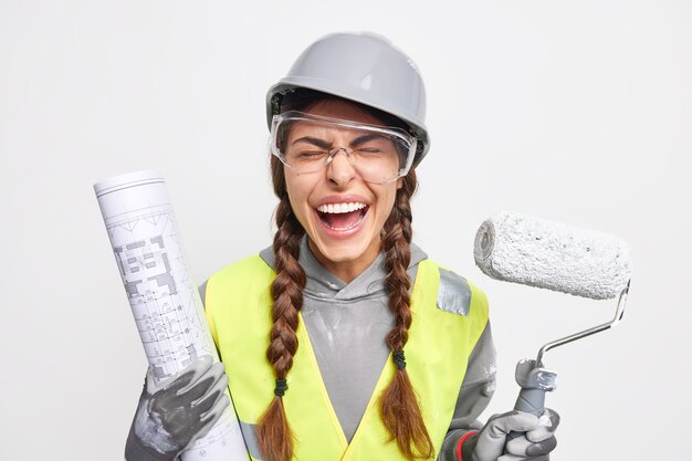 Concept d'entretien et d'occupation. Une ingénieure ravie pose avec un plan et un rouleau à peinture vêtus de vêtements de sécurité occupés à reconstruire sur un chantier de construction isolé sur un mur blanc