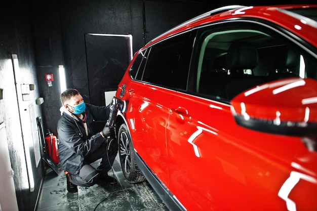 Concept d'esthétique automobile Homme en masque facial avec polisseuse orbitale dans un atelier de réparation polissant une voiture suv orange