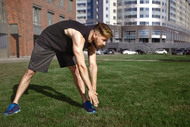 Concept d'été, d'activité, de remise en forme et de loisirs. Musclé jeune homme mal rasé européen en vêtements de sport noirs et chaussures de course se penchant vers l'orteil gauche, étirement des jambes tout en exerçant à l'extérieur