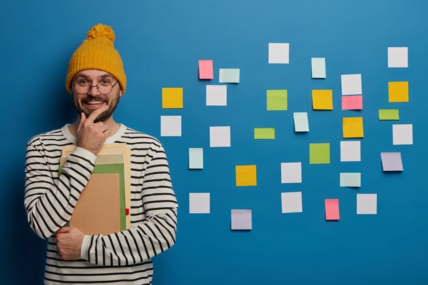 Concept d'étude, d'apprentissage et d'éducation. Joyeux étudiant masculin tient le menton, sourit joyeusement, se tient avec un cahier et un manuel, vêtu de vêtements à la mode, utilise des notes autocollantes sur le mur bleu