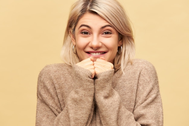 Concept d'excitation, de plaisir et d'attitude positive. Image isolée de belle jolie fille élégante portant un pull confortable serrant les poings sur son visage et souriant, anticipant quelque chose d'étonnant