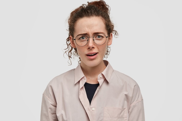 Concept d'expressions faciales négatives. Malheureuse femme aux taches de rousseur indignée aux cheveux bouclés, fronce les sourcils avec mécontentement, porte une chemise surdimensionnée beige, entend quelque chose de désagréable de l'interlocuteur