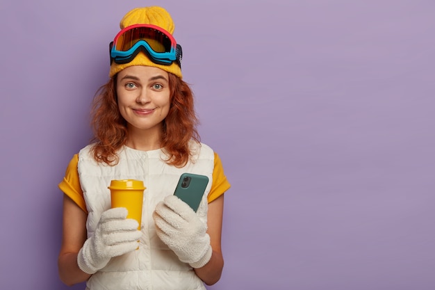 Concept extrême de sports d'hiver, de loisirs et de technologie. Une femme rousse heureuse tient du café à emporter et un téléphone mobile moderne, étant une skieuse professionnelle, publie des photos sur les réseaux sociaux.
