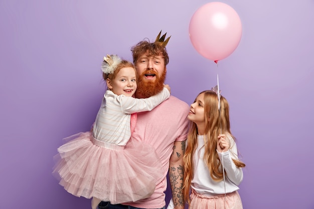 Concept de famille, parentalité et vacances. Père drôle divertit les enfants, porte une petite fille sur les mains, décore la pièce avant la fête, porte des vêtements de fête, isolés sur un mur violet.