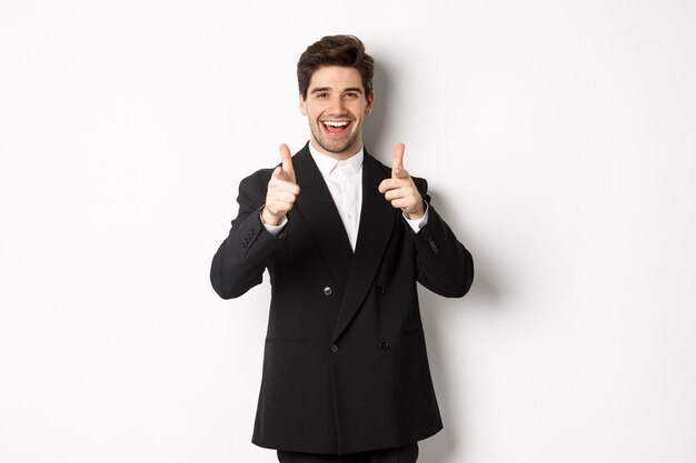 Concept de fête du nouvel an, de célébration et de style de vie. Mec beau et réussi en costume noir, pointant du doigt la caméra et vous félicitant, debout sur fond blanc.