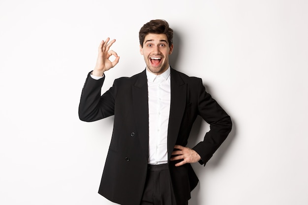 Concept de fête du nouvel an, de célébration et de style de vie. Portrait d'un beau mec heureux en costume noir, loue quelque chose de bien, montre bien son approbation, debout sur fond blanc.