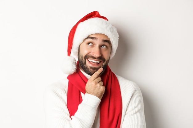 Concept de fête du nouvel an et de vacances d'hiver. Gros plan sur un bel homme barbu à la recherche d'une liste de cadeaux de Noël réfléchie, en bonnet de Noel, fond blanc