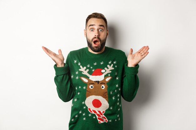 Concept de fête du nouvel an et de vacances d'hiver. Mec barbu surpris haletant étonné, debout dans un pull de Noël, fond blanc