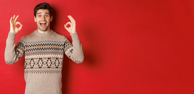 Concept de fête de noël, vacances d'hiver et style de vie. Portrait d'un bel homme en pull, l'air étonné et montrant des signes corrects, garantir ou recommander quelque chose de bon, fond rouge.