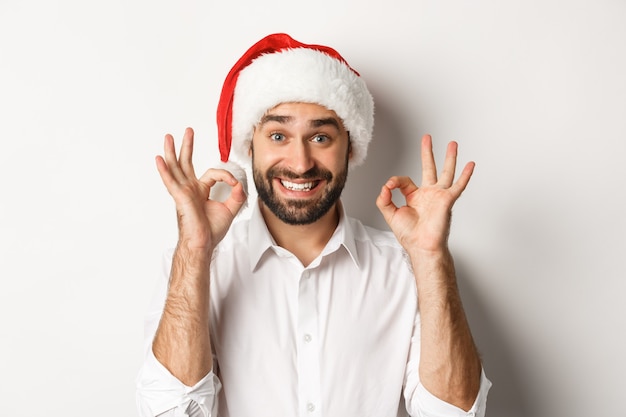 Concept de fête, vacances d'hiver et célébration. Homme joyeux appréciant Noël et montrant un signe correct, souriant satisfait, portant un bonnet de Noel