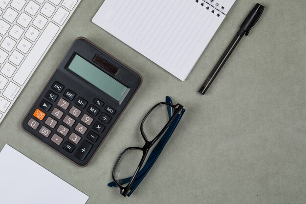 Concept financier avec ordinateur portable, papier, stylo, calculatrice, clavier, lunettes sur fond plat gris.