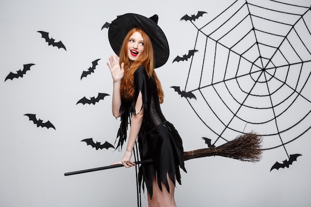 Concept d'Halloween, une sorcière élégante et heureuse aime jouer avec un balai fête d'halloween sur un mur gris