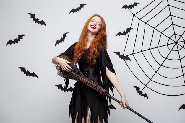 Concept d'Halloween, une sorcière élégante et heureuse aime jouer avec un balai fête d'halloween sur un mur gris
