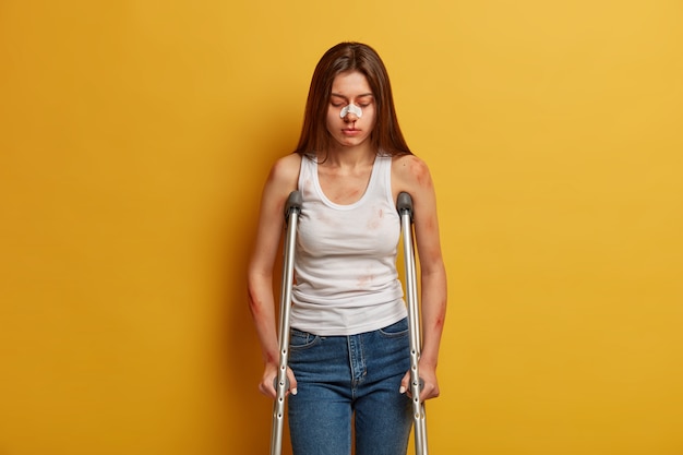 Concept de handicap et de problèmes de santé. Une femme malheureuse a subi un grave traumatisme lors d'un accident, utilise l'aide à la mobilité, fait ses premiers pas après la chirurgie, regarde vers le bas, porte du plâtre sur le nez, pose à l'intérieur