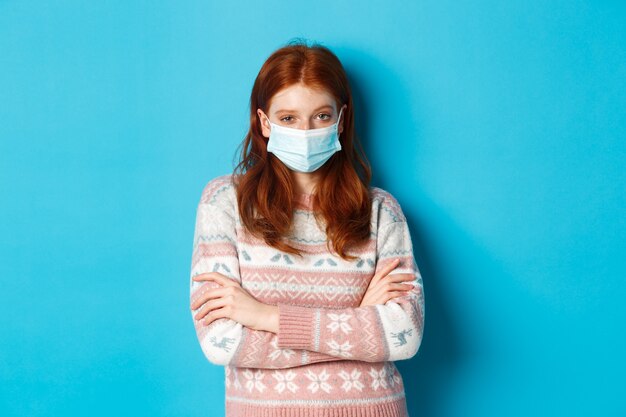 Concept d'hiver, de covid-19 et de quarantaine. Fille rousse sceptique en pull et masque médical, bras croisés sur la poitrine et regardant avec incrédulité, debout sur fond bleu