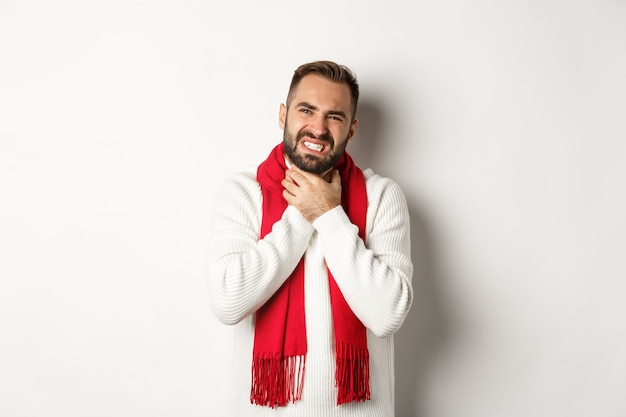 Concept hiver, covid-19 et santé. Homme malade se plaignant d'un mal de gorge, touchant le cou et grimaçant de douleur, debout dans un pull et une écharpe rouge, fond blanc