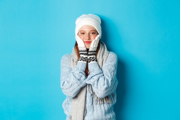 Concept d'hiver et de vacances. Jolie fille en bonnet blanc, pull et gants, serrant les joues et souriant heureux, échauffement après le froid à l'extérieur, debout sur fond bleu