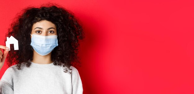 Photo gratuite concept immobilier et covid femme excitée dans un masque médical montrant une petite découpe de maison en papier standin