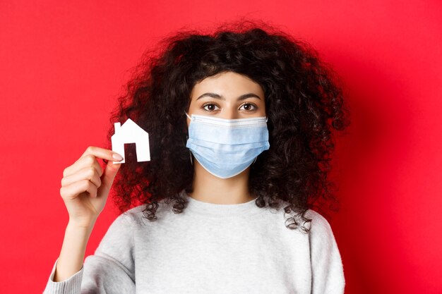 Concept immobilier et covid femme excitée dans un masque médical montrant une petite maison en papier se tenant debout...