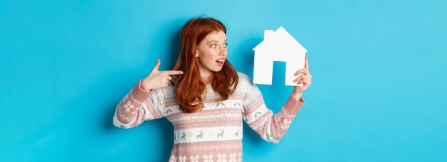 Photo gratuite concept immobilier femme rousse excitée aux cheveux rouges pointant et regardant le modèle de maison en papier s