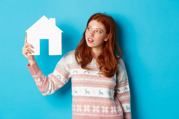 Concept immobilier. Image d'une jolie fille rousse à la recherche d'un modèle de maison en papier, pensant à acheter une propriété, debout en pull sur fond bleu.