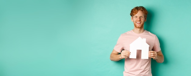 Photo gratuite concept immobilier jeune homme aux cheveux rouges portant un t-shirt montrant la découpe de la maison en papier et souriant ha