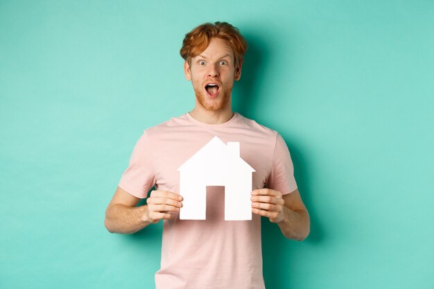 Concept immobilier. Rousse excité avec barbe, montrant la découpe de la maison en papier et le souffle coupé, regardant fasciné par la caméra, debout sur fond de menthe