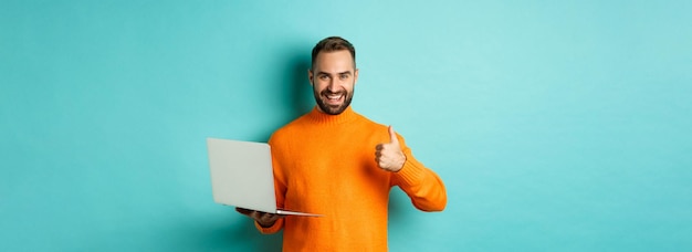 Photo gratuite concept indépendant et technologique homme chanceux en pull orange montrant le pouce vers le haut tout en travaillant avec les genoux