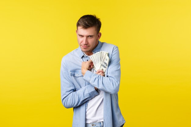 Concept d'investissement, de shopping et de finance. Un gars blond drôle et gourmand serrant de l'argent et regardant la caméra, montrant un désir intense de garder de l'argent, une part réticente, un fond jaune debout.