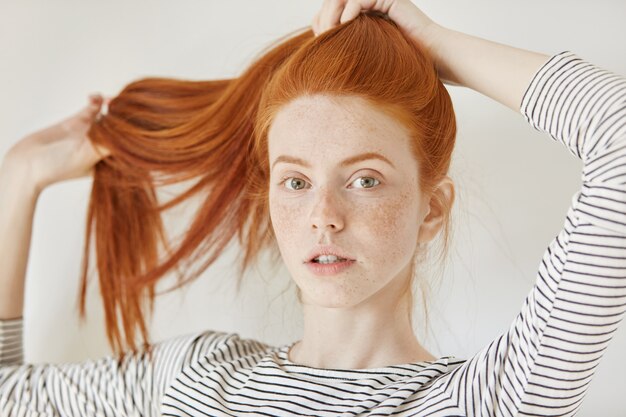 Concept de jeunesse, âge tendre et mode de vie. Jeune femme à la mode avec des taches de rousseur attachant ses beaux cheveux roux en queue de cheval