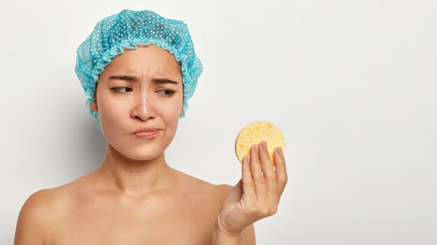 Concept de jeunesse, de soins, de nettoyage et de soins de la peau. Insatisfaite jeune femme chinoise regarde malheureusement l'éponge cosmétique, enlève le maquillage du visage, porte un couvre-chef de protection bleu