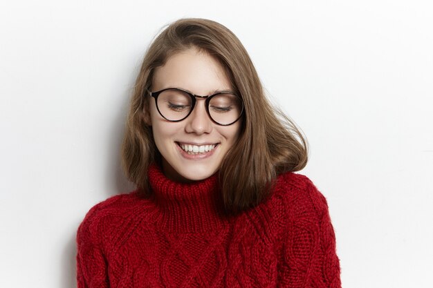 Concept de joie et de bonheur. Photo de mystérieuse jeune femme de race blanche ludique portant des lunettes transparentes et élégant pull à col roulé marron, regardant vers le bas avec un sourire timide, être embarrassé