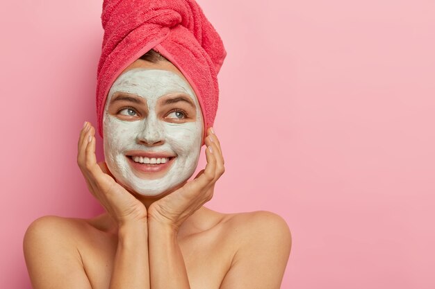 Concept de jour de spa. Belle femme heureuse sourit positivement, montre les dents, touche doucement le visage, applique un masque de beauté pour le rajeunissement et le nettoyage des pores, a le corps nu, regarde de côté contre le mur rose