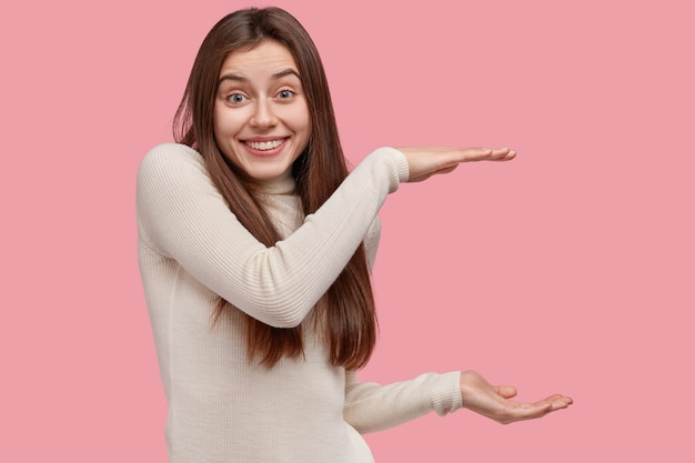 Concept De Langage Corporel. Heureux Femme De Race Blanche Montre La Taille De La Boîte à Deux Mains, Sourit Volontiers, Porte Un Pull Blanc Décontracté, Isolé Sur Un Mur Rose