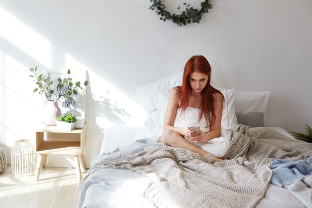 Concept de literie, de loisirs et de technologies modernes. Plan intérieur d'une belle jeune femme rousse restant au lit pendant la journée, surfant sur Internet sur un téléphone portable, envoyant des messages à des amis via les réseaux sociaux