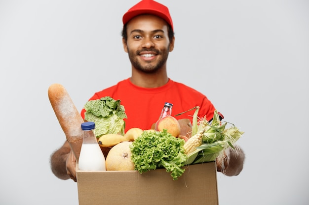 Concept de livraison - Handsome African American Delivery Man portant une boîte à colis d&#39;épicerie et des boissons au magasin. Isolé sur fond de studio gris. Espace de copie.