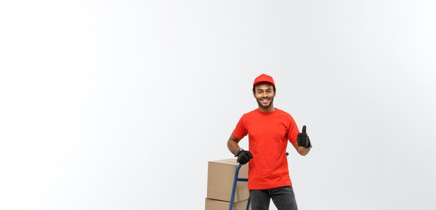 Concept de livraison Portrait d'un beau livreur afro-américain ou d'un coursier poussant un camion à main avec une pile de boîtes isolé sur fond de studio gris espace de copie