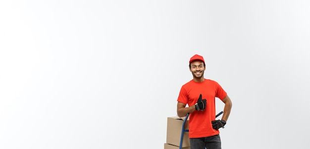 Concept de livraison Portrait d'un beau livreur afro-américain ou d'un coursier poussant un camion à main avec une pile de boîtes isolé sur fond de studio gris espace de copie