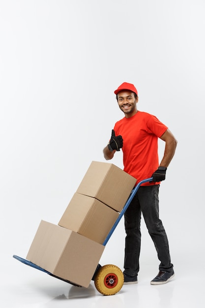 Concept de livraison - Portrait de Handsome African American Delivery Man ou de messager poussant un camion à main avec pile de boîtes. Isolé sur fond de studio gris. Espace de copie.