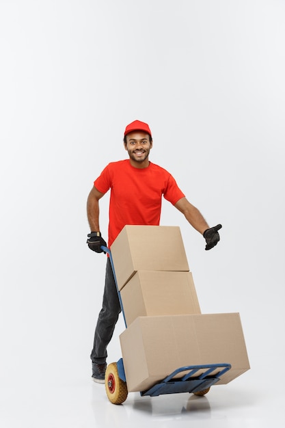 Concept de livraison - Portrait de Handsome African American Delivery Man ou de messager poussant un camion à main avec pile de boîtes. Isolé sur fond de studio gris. Espace de copie.