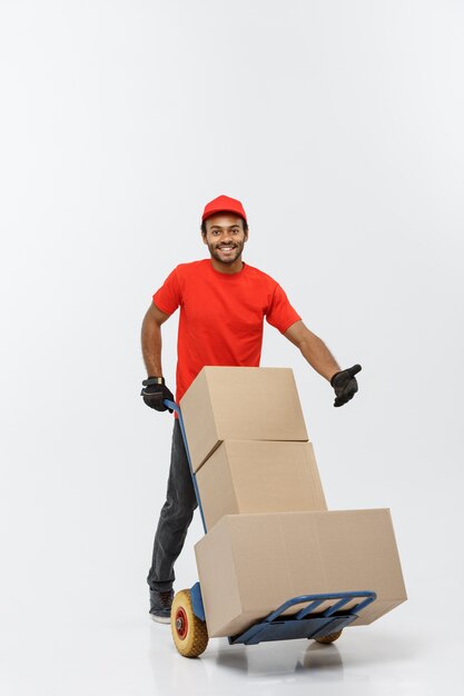 Concept de livraison - Portrait de Handsome African American Delivery Man ou de messager poussant un camion à main avec pile de boîtes. Isolé sur fond de studio gris. Espace de copie.