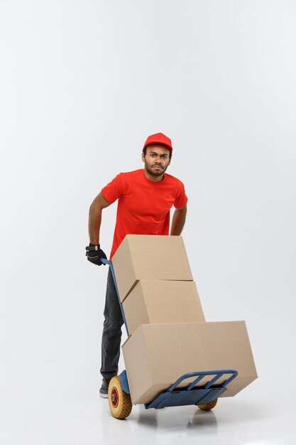 Concept de livraison - Portrait de Handsome African American Delivery Man ou de messager poussant un camion à main avec pile de boîtes. Isolé sur fond de studio gris. Espace de copie.