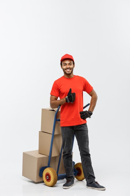 Concept de livraison - Portrait de Handsome African American Delivery Man ou de messager poussant un camion à main avec pile de boîtes. Isolé sur fond de studio gris. Espace de copie.