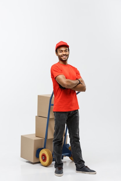 Concept de livraison - Portrait de Handsome African American Delivery Man ou de messager poussant un camion à main avec pile de boîtes. Isolé sur fond de studio gris. Espace de copie.